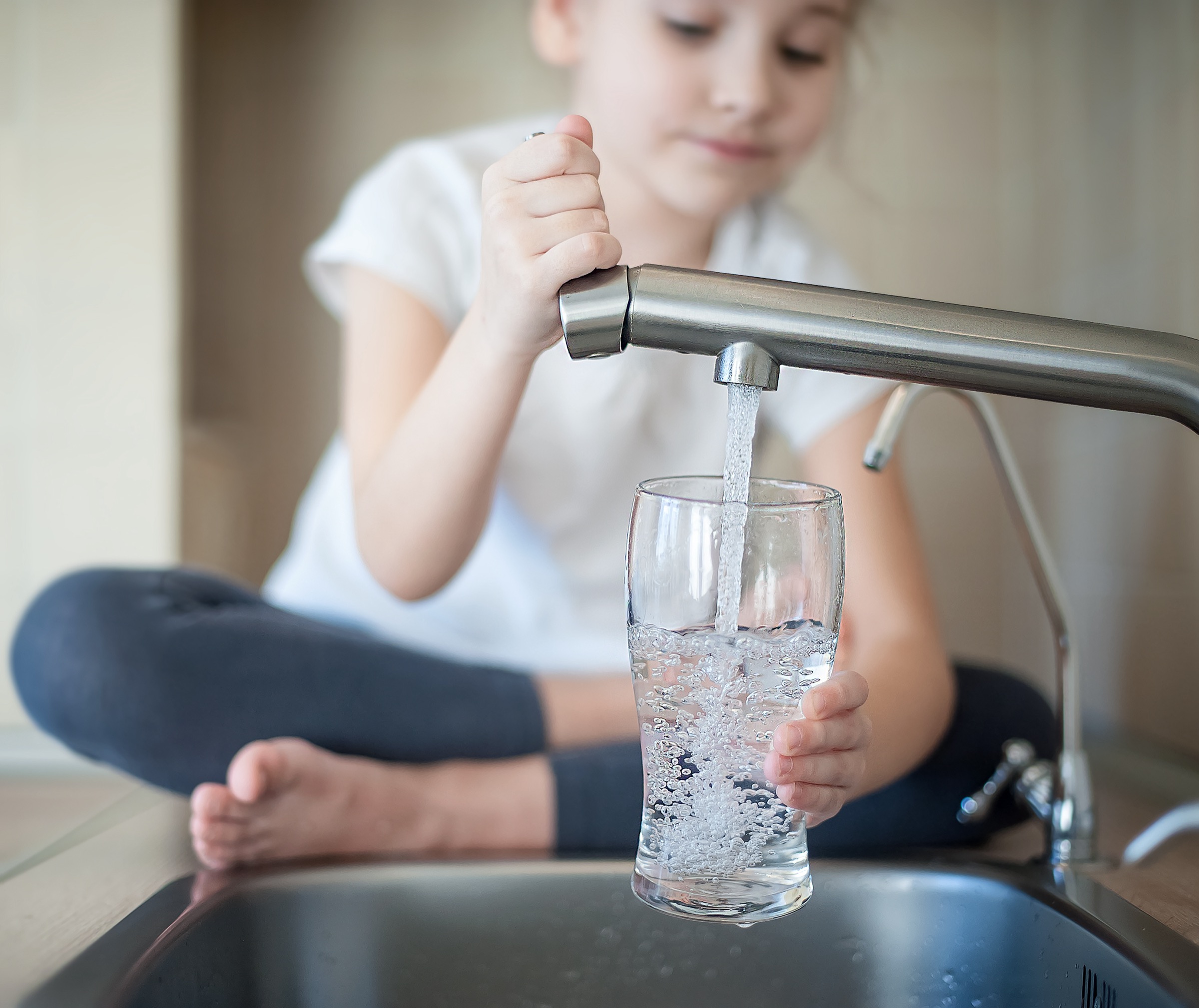 Little girl opens a water tap with her hand holding a transparent glass. 